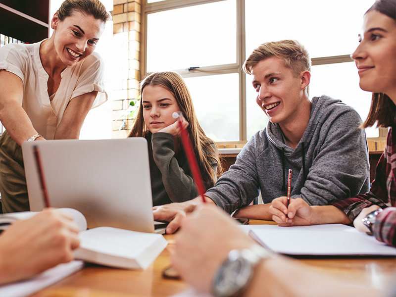 Schülerinnen und Schüler sitzen gemeinsam an einem Tisch und blicken auf ein Tablet. Eine Lehrerin zeigt ihnen etwas auf dem Tablet.