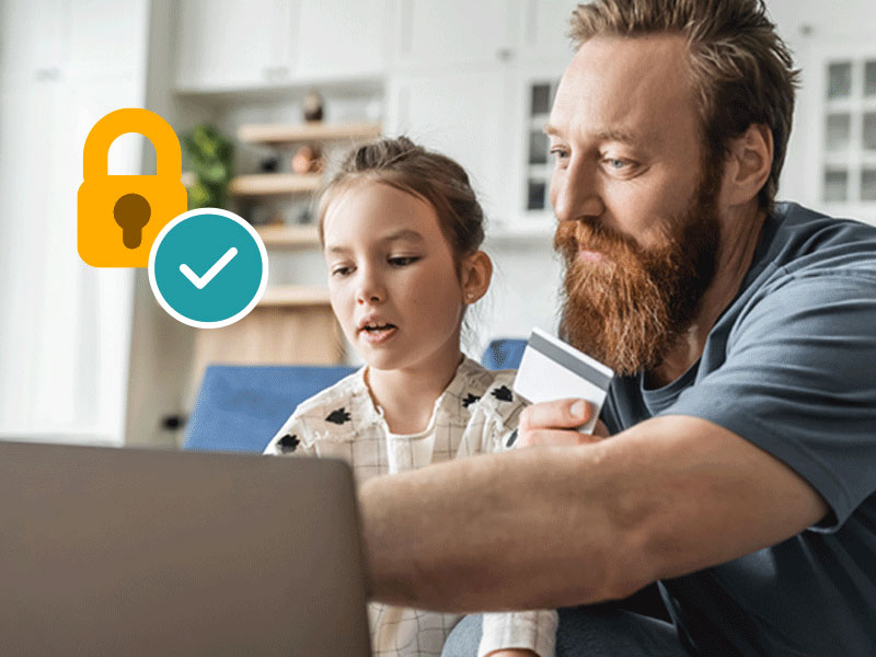 Ein Mann mit einem rötlichen Arm hat eine Bankkarte in der Hand und zeigt auf einen Laptop. Neben ihm sitzt seine Tochter und schaut ebenfalls auf den Laptop.