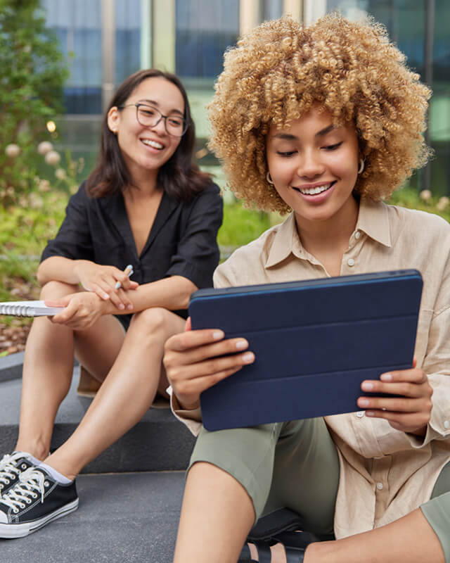 Zwei Frauen sitzen draußen und schauen auf ein Tablet.